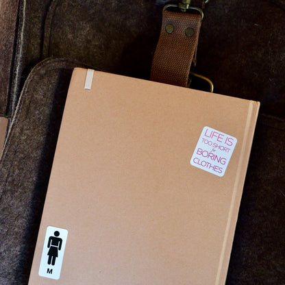 Brown journal on top of a dark brown messenger bag. Overhead view. Journal has two stickers: 'Life is too short for boring clothes' and a men's washroom / restroom symbol but with a skirt instead of pants, and 'M' below to signify male or men.